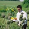 A man carrying a basket of vegetables walks past large flowers in a field