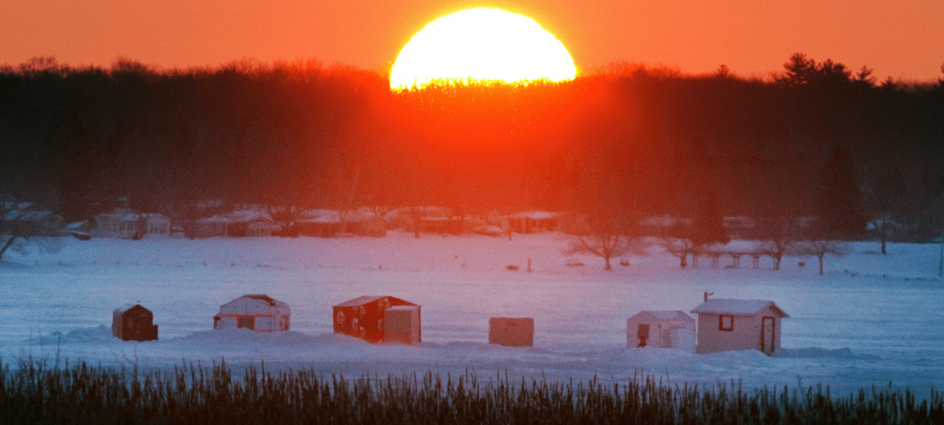 A brilliant orange sun on the horizon illuminates a row of ice fishing huts