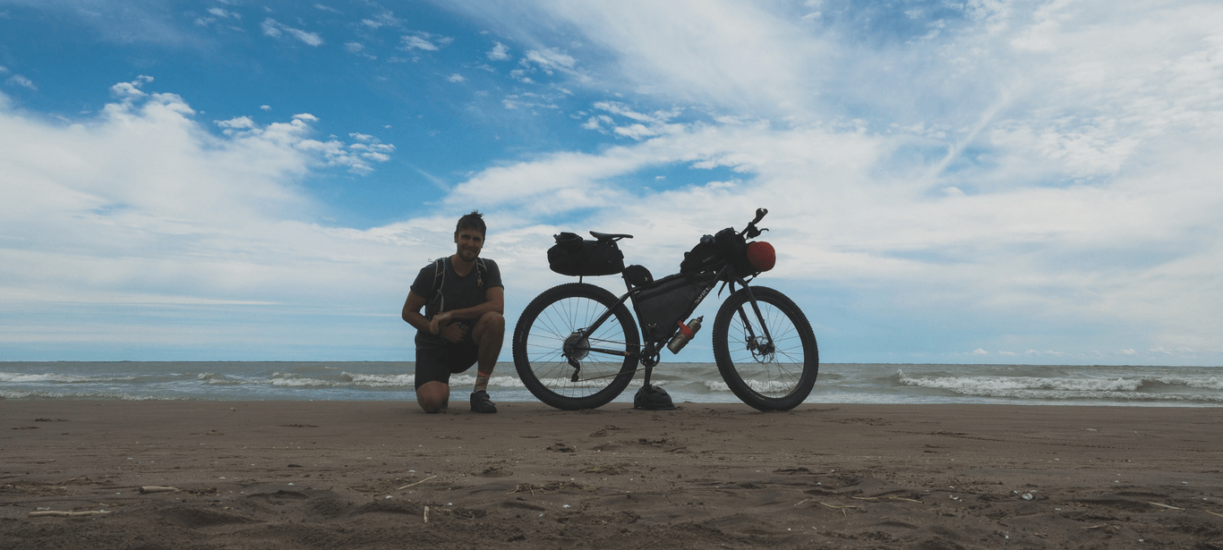 A cyclist kneels beside his bike on a beach
