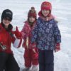 A woman holding a fishing rod kneels and gives the thumb up beside two smiling girls standing on a frozen lake