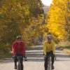 Two men ride bikes side by side down a backroad in fall