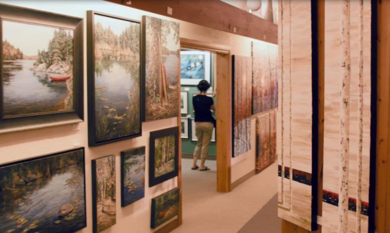 Interior of Gallery on the Lake, showing a wall of landscape paintings with a woman looking at painting in the background