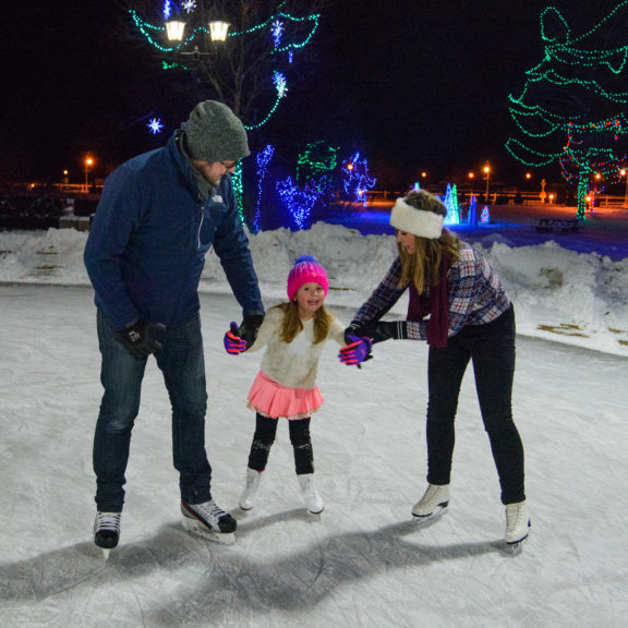 A couple holds a little girl's hands while she skates outdoors in Cobourg, with Christmas lights in the background