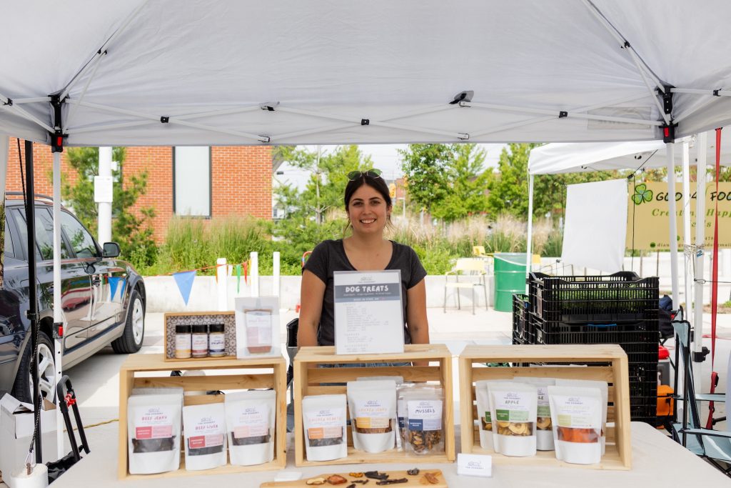 A smiling woman poses at a farm stand that sells a variety of boutique dog treats