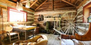 Interior of a log cabin, with large wagon wheel, crib, table, and hanging pots