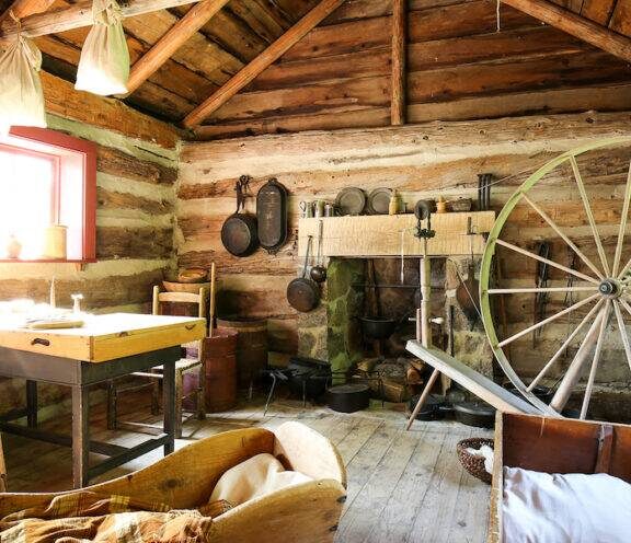 Interior of a log cabin, with large wagon wheel, crib, table, and hanging pots