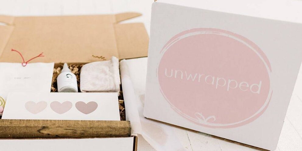 A pink and white box branded Unwrapped, with the lid set to the side and the contents showing, which include a card with hearts, a cloth bag, and a small bottle