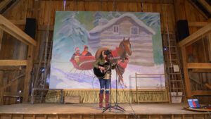 A woman on a stage in a barn plays guitar in front of a mural of a horsedrawn sleigh