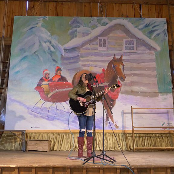 A woman on a stage in a barn plays guitar in front of a mural of a horsedrawn sleigh