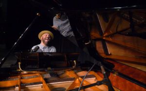 Beau Dixon performs on a grand piano, and is partly reflected in the glossy raised lid
