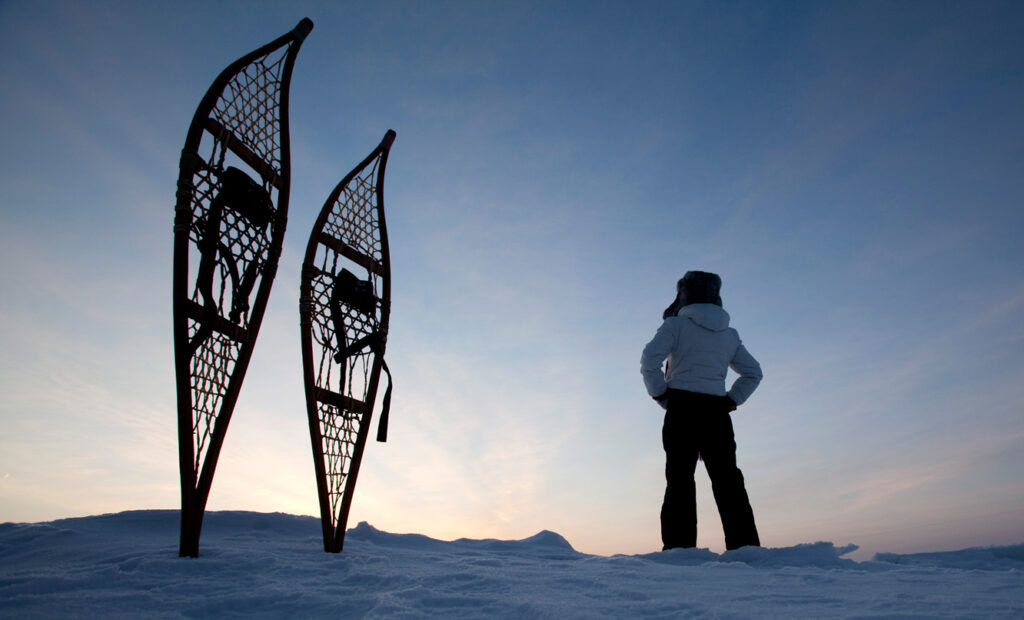 A person looks out at the sunset with satisfaction, with snowshoes propped in the foreground