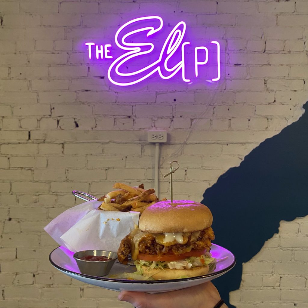 Hand holds a burger under a sign reading The El (P)