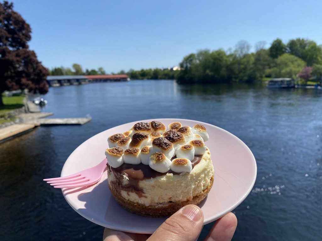 Hand holds a mini-cheesecake against a lakeside backdrop