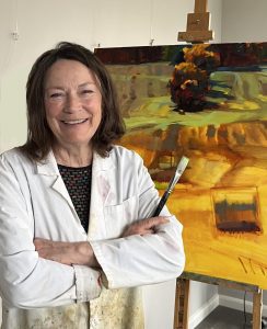 Painting Jane Robertson stands smiling beside one of her agricultural landscape paintings