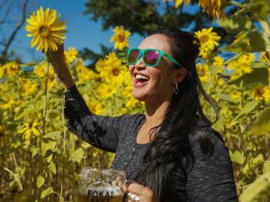 A woman in sunglasses holding a pint glasses wanders a sunflower field