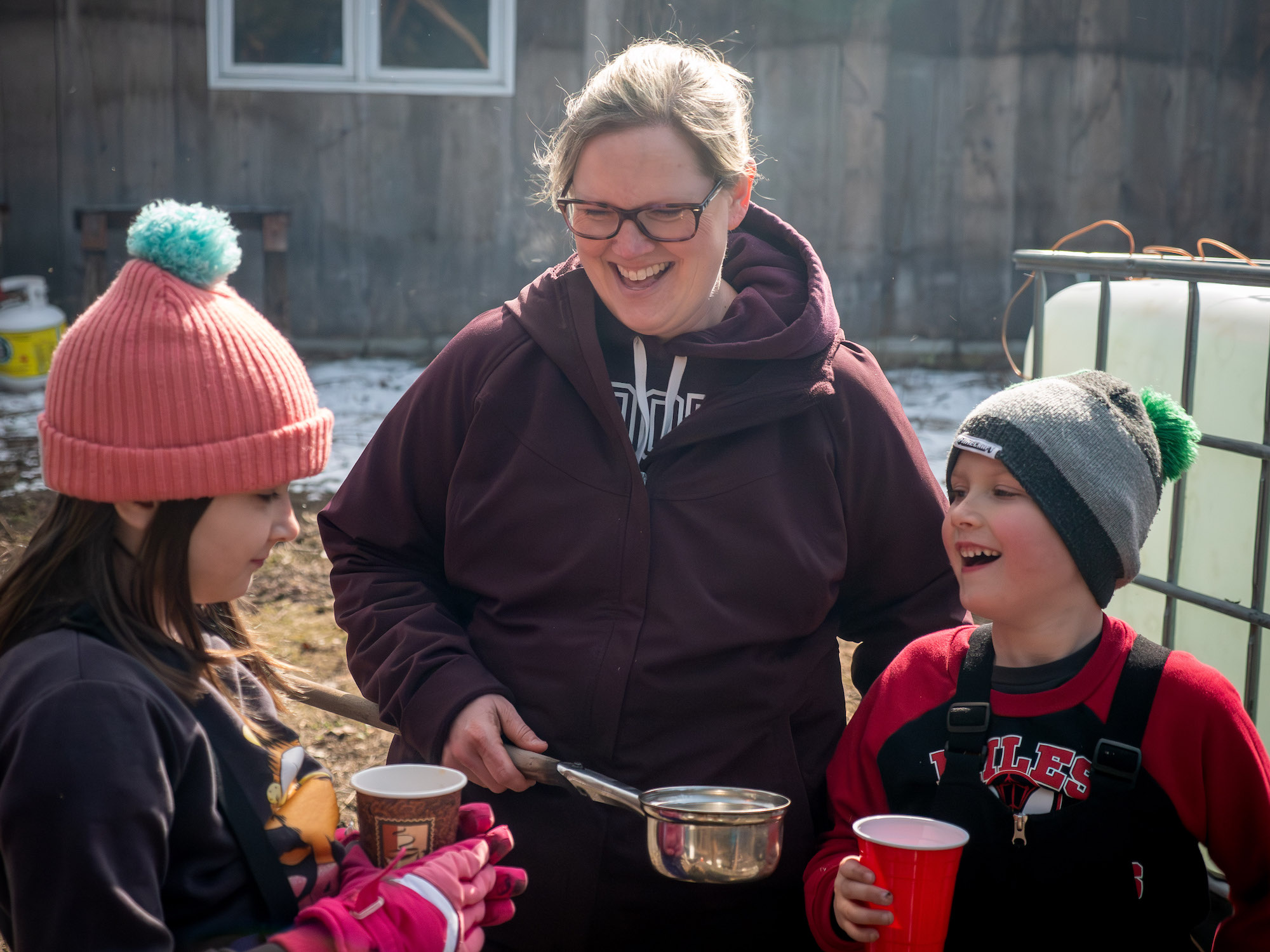 A smiling woman with two children on either side, each holding a maple syrup sample