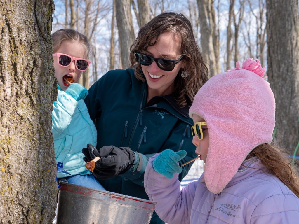 A woman and two children inspect a maple sap bucket on a tree