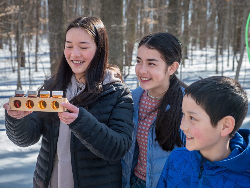 Three young people smile enthusiastically while one of them holds a selection of maple syrup samples