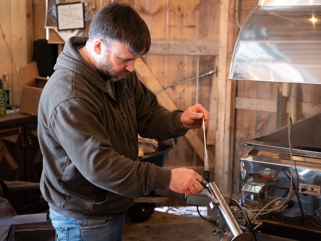 A man standing beside an evaporator tests the progress of maple syrup