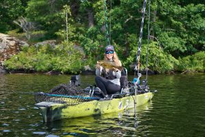 A woman in a yellow kayak in a river holds a fish towards the camera