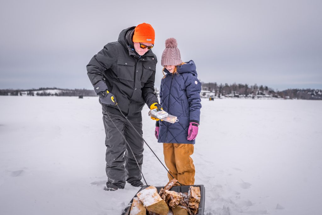 A man in a black winter coat and orange toque holds fishing tackle and a sled of firewood; a girl in a blue coat and toque looks on 