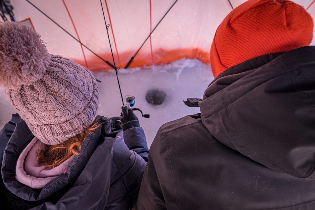 Over-the-shoulder shot of girl in grey toque and man in orange toque around ice fishing hole