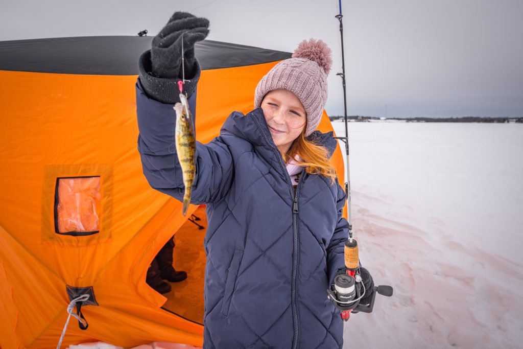 A girl in winter gear outside an orange ice hut holds up a recently-caught perch