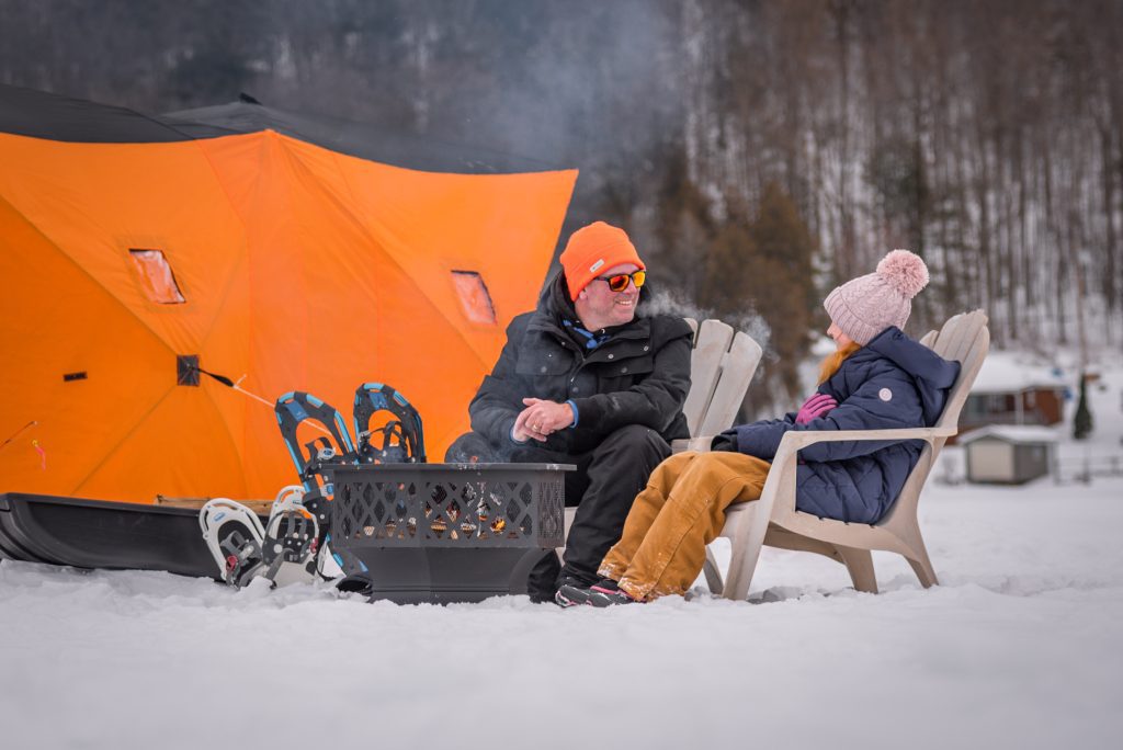 A man and a girl in winter gear sit around a firepit outside and orange ice hut