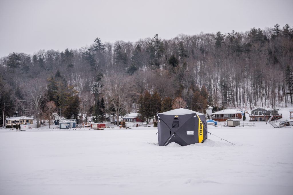 A black and yellow ice hut, with cottages and a wooded ridge in the background