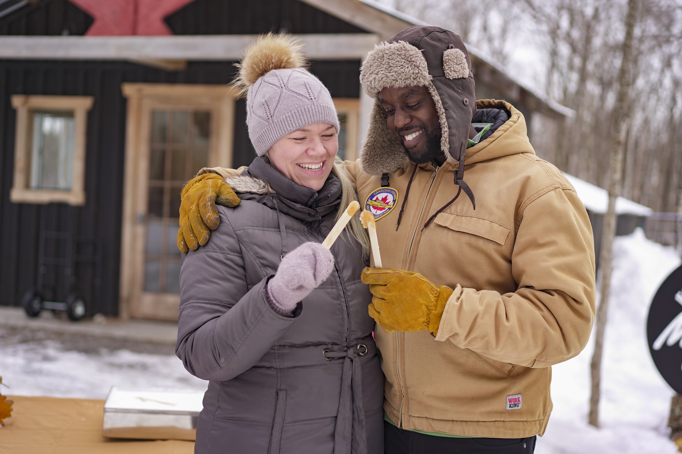 A smiling man and woman in winter gear stand close together holding sticks of maple taffy