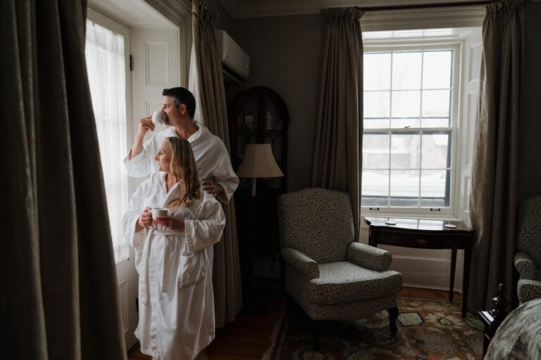 A man and woman in bathrobes look out the window while sipping from ceramic mugs