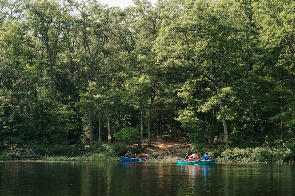 Two canoes carrying three people each arrive at the banks of a  shady wooded campsite