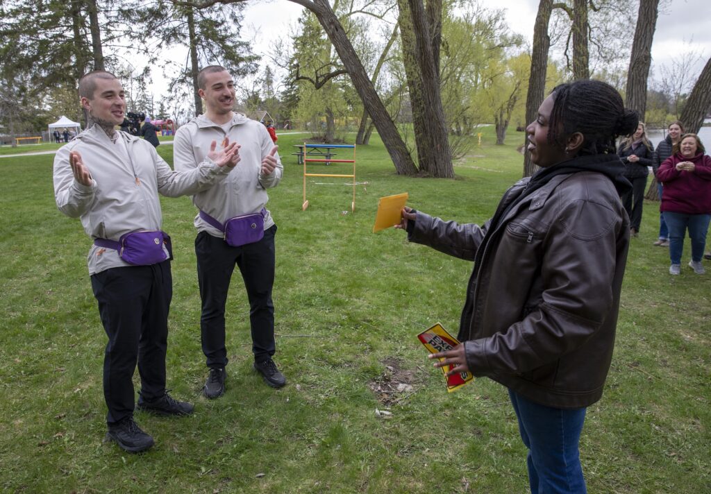Two smiling men in a park accept an envelope from a person in a leather jacket