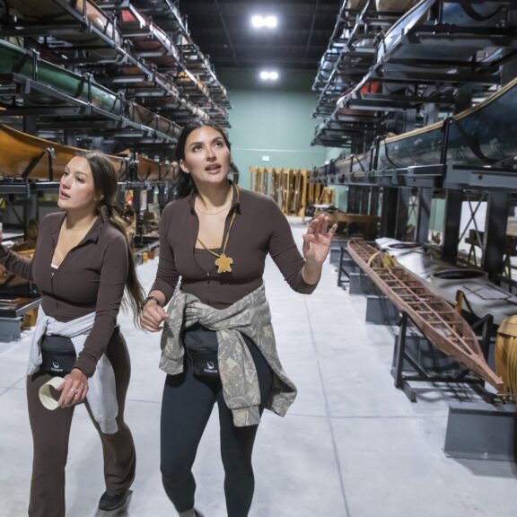 Two women walk through a museum collection hall full of canoes