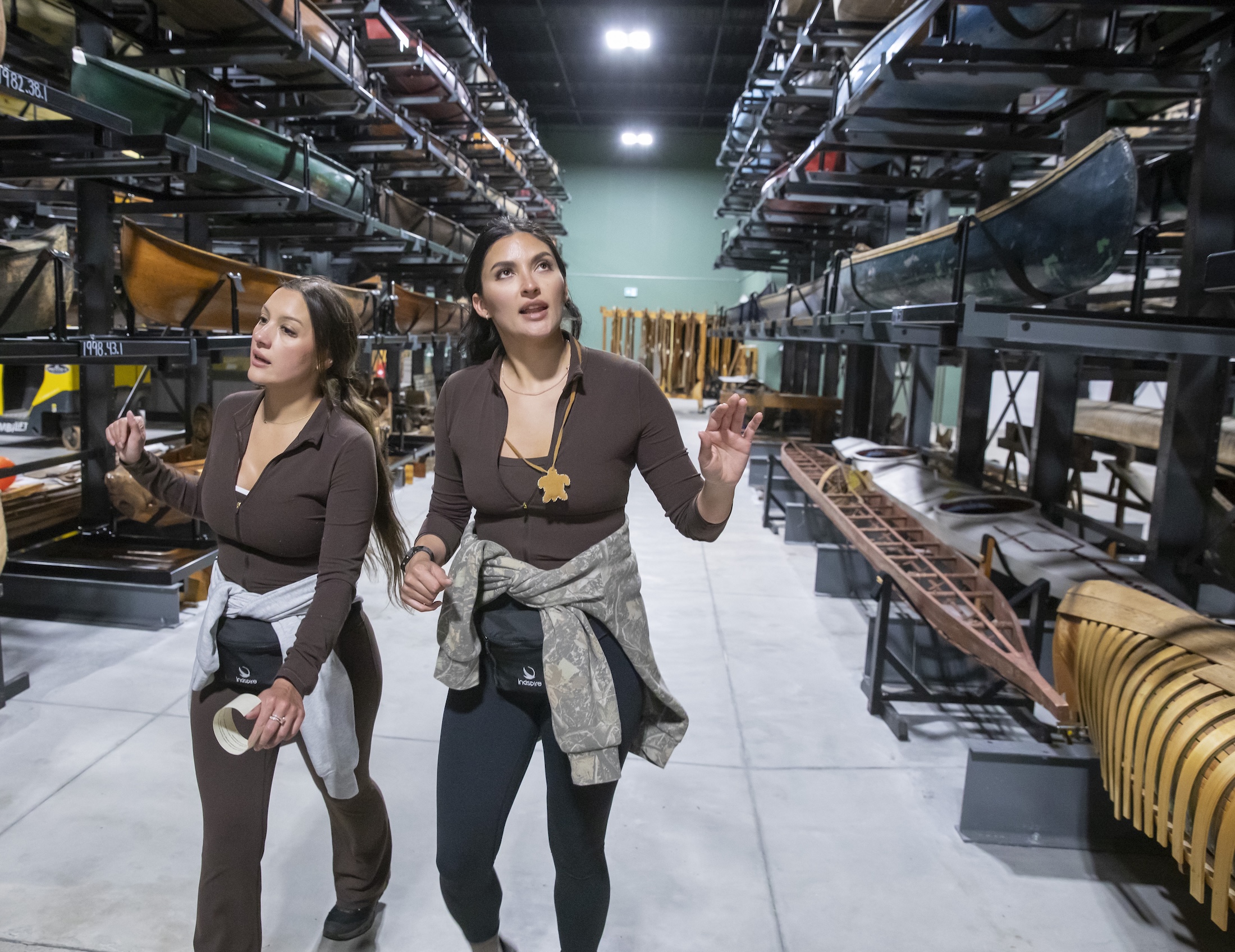 Two women walk through a museum collection hall full of canoes