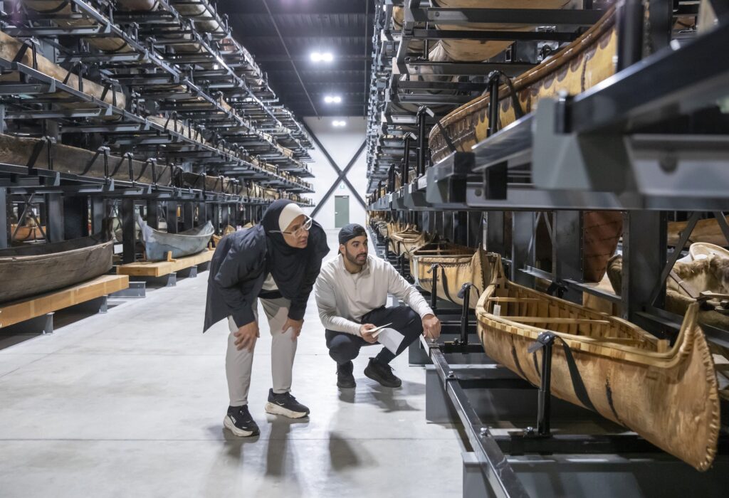 A woman and a man bend in to look closely at a canoe, while standing among a large collection of canoes