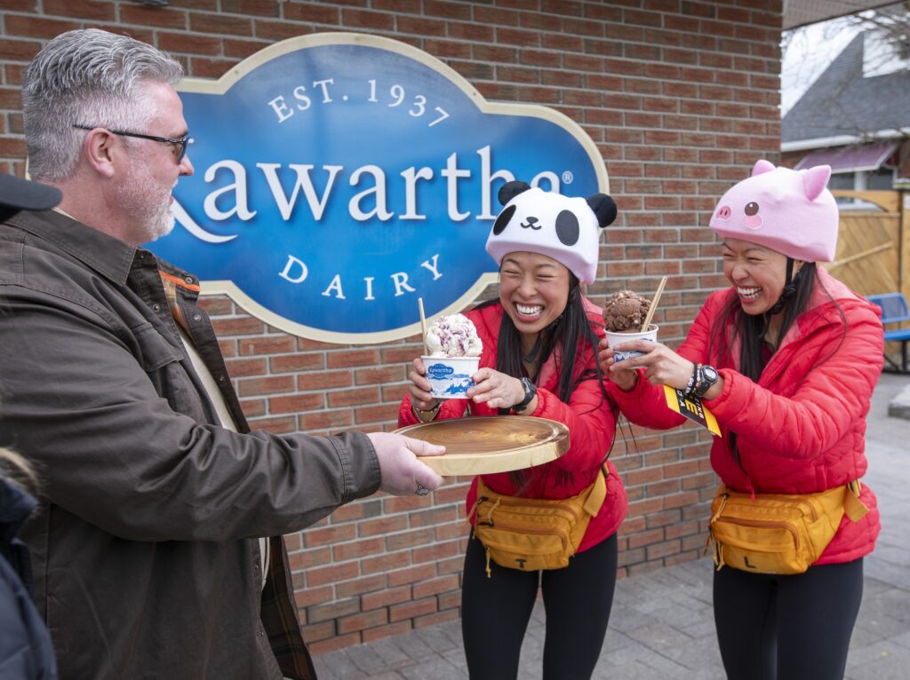 Two smiling women in red jackets accept cups of ice cream from a man with a wooden serving tray; a Kawartha Dairy sign hangs in the background